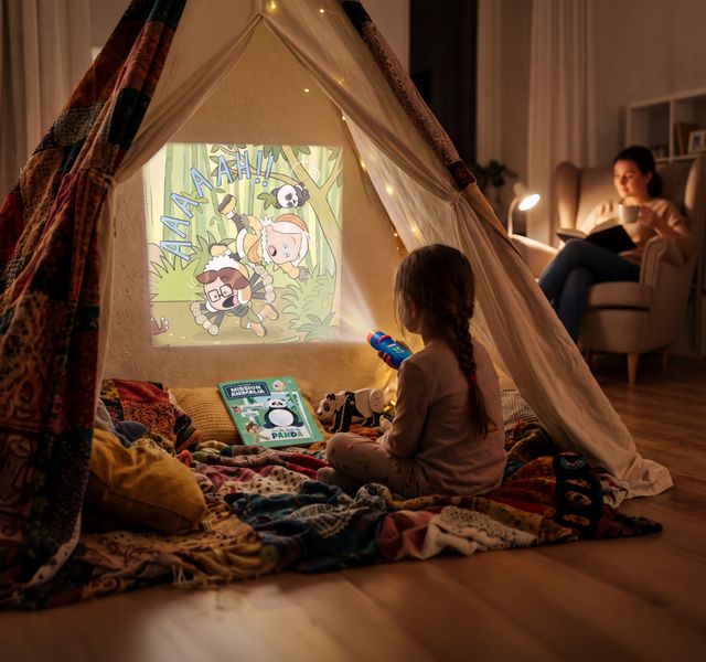 Sous sa tente, une petite fille projette une histoire de Mission Animalia sur la toile avec sa lampe à histoire ; derrière elle, sa mère lit avec un café.